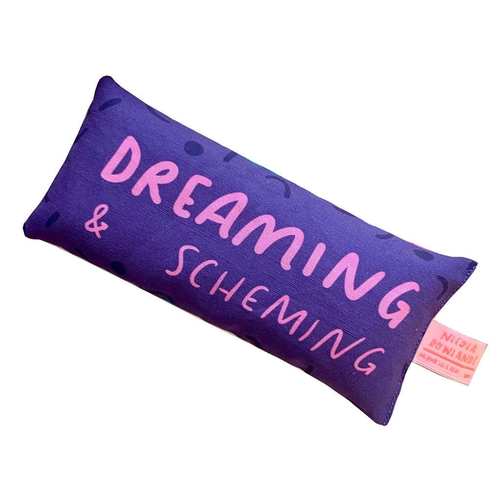 Nicola Rowlands Dreaming & Scheming lavender scented weighted eye pillow with pink lettering on a purple background.