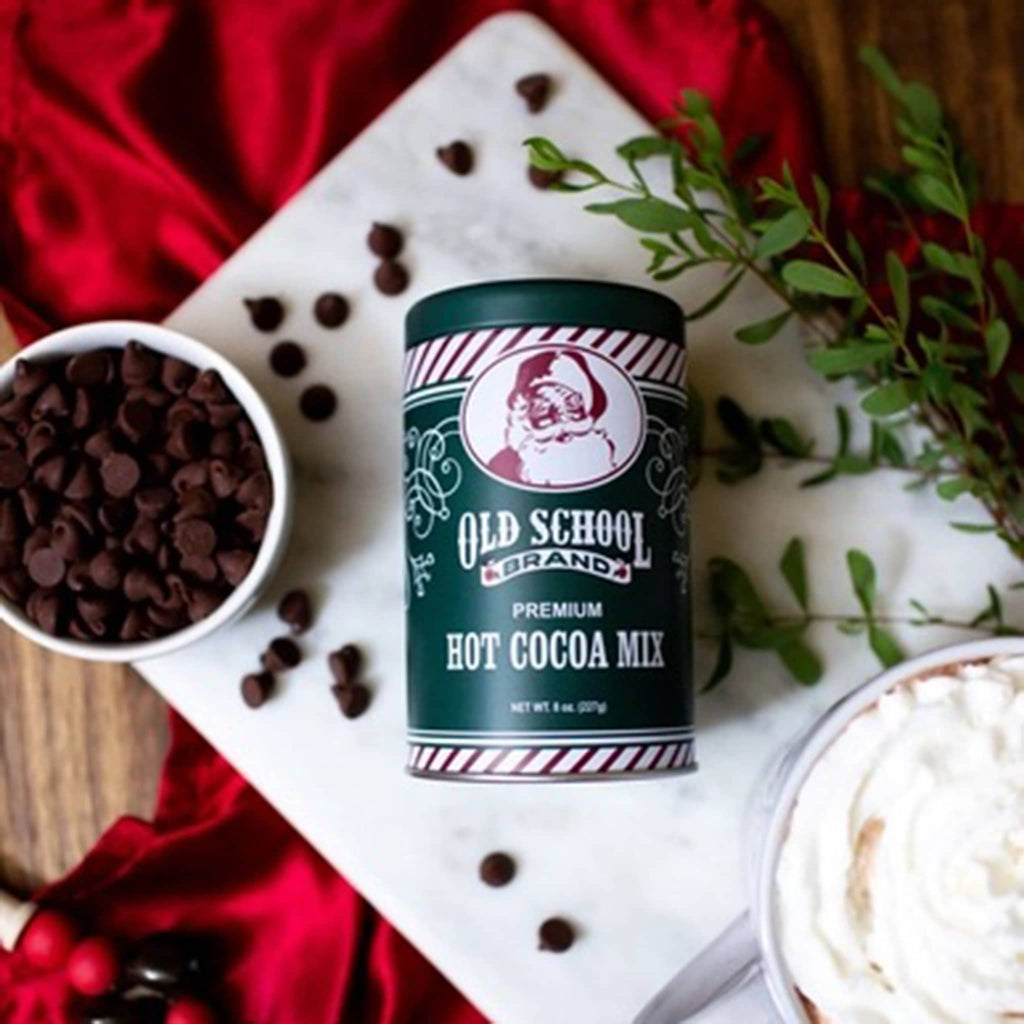 Old School Brand hot cocoa mix in green and white tin canister with santa illustration, on marble with chocolate chips.