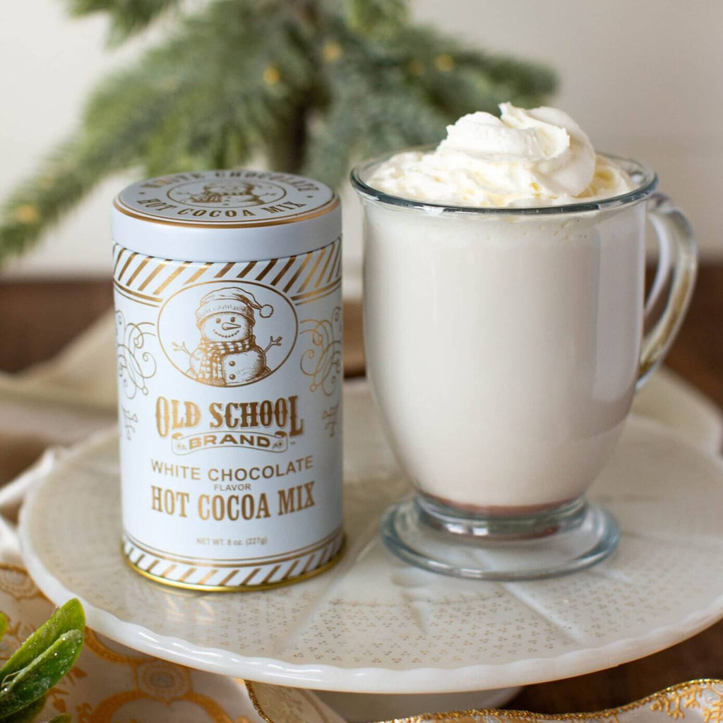 Old School Brand white chocolate hot cocoa mix in a white tin canister with gold lettering and snowman illustration, beside a mug of prepared hot chocolate.