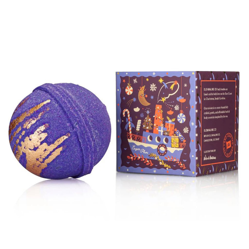 Purple bath bomb with gold accents next to its decorative box on a white background.