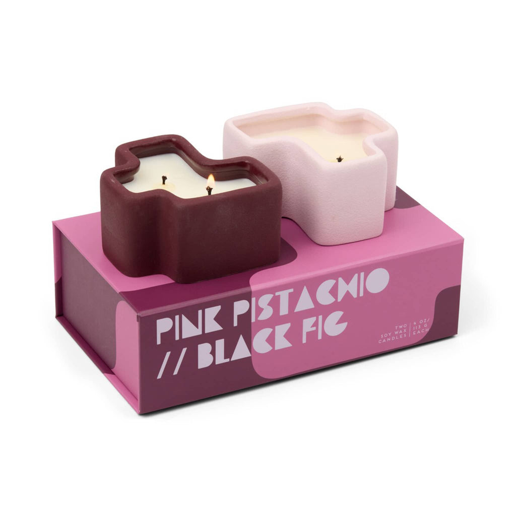 Two candles in decorative puzzle piece shaped holders on a pink and burgundy box labeled"Pink Pistachio and Black Fig".