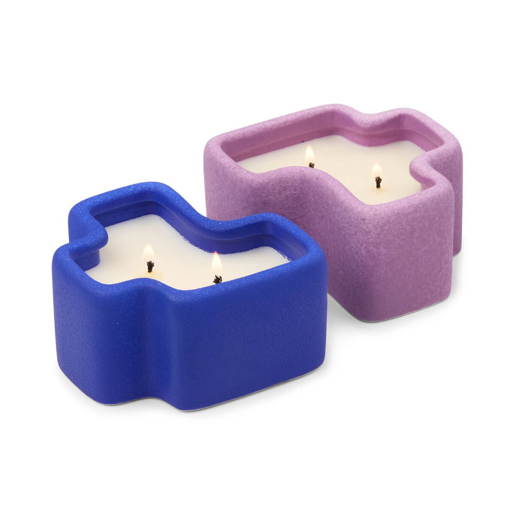 Two ceramic candle holders in cobalt blue and lilac, shaped like puzzle pieces with double wicks lit, on a white background.