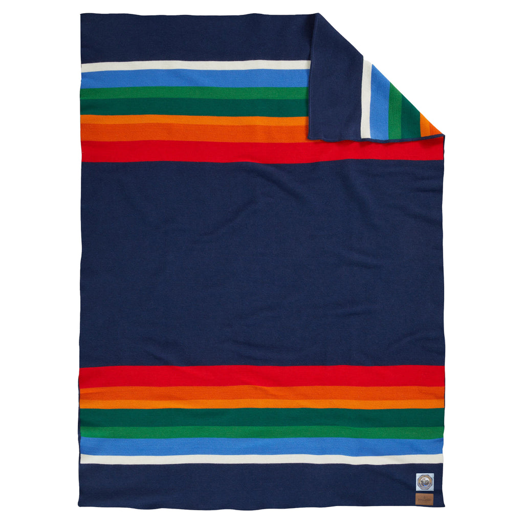 Pendleton Crater Lake National Park Throw Blanket in navy with white, blue, green, orange and red stripes at both ends, full blanket shown with a corner folded back.