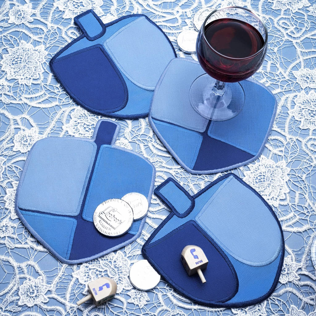 Set of four blue linen dreidel napkins with unique designs on a lace tablecloth with a glass of red wine and gelt.