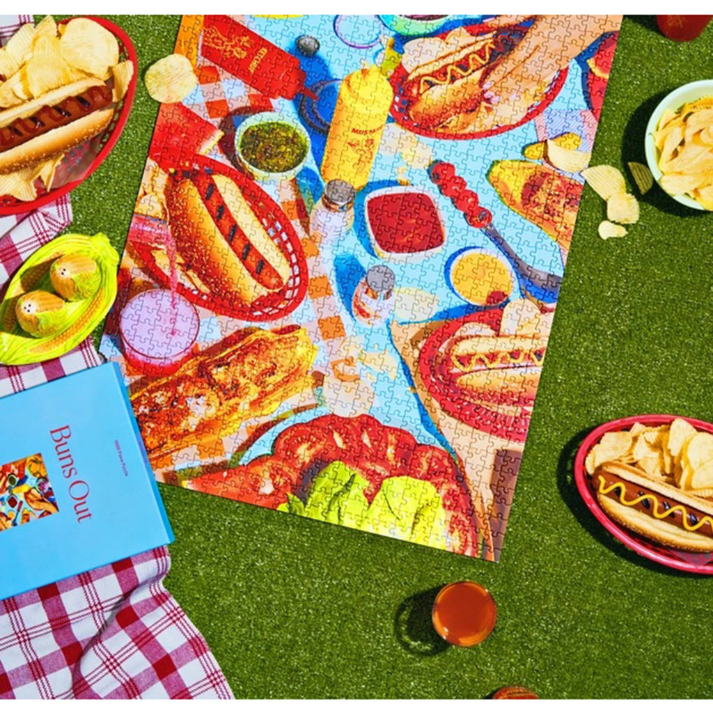 Piecework Puzzles 1000 Piece Buns Out jigsaw puzzle in light blue box packaging beside finished puzzle surrounded by hot dogs and chips on fake grass backdrop.