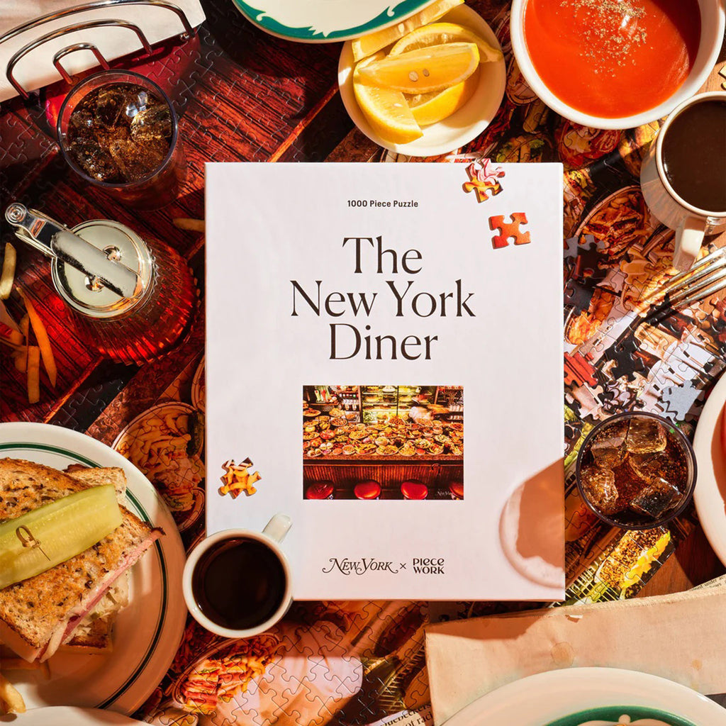 Piecework Puzzles x New York Magazine 1000 piece The New York Diner jigsaw puzzle in white box packaging, front view with small puzzle image. Shown surrounded by diner food on the partially completed puzzle.