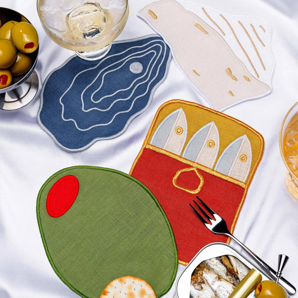 Piecework hors d'oeuvres themed linen cocktail napkins with olives and a tin of sardines, on a white tablecloth.
