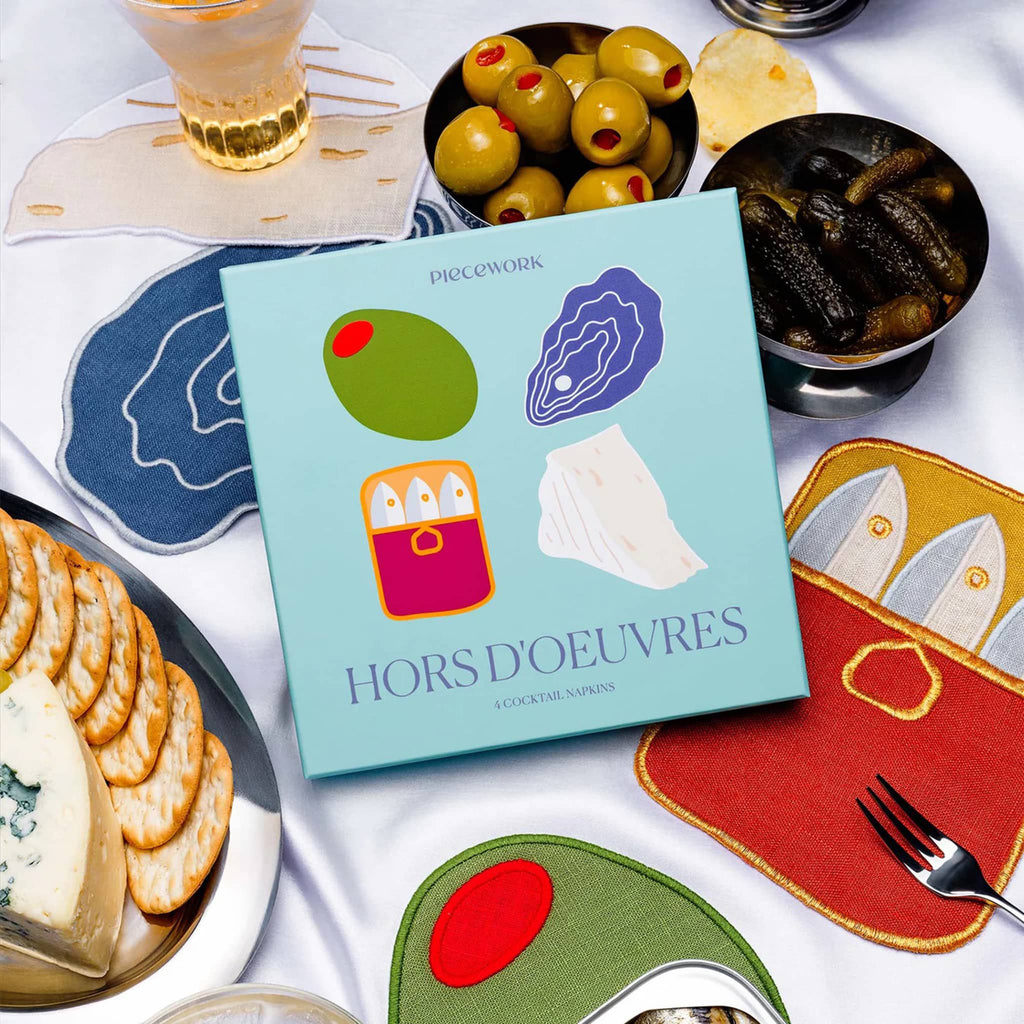 Piecework hors d'oeuvres themed linen cocktail napkins with box packaging and food props on a tablecloth.