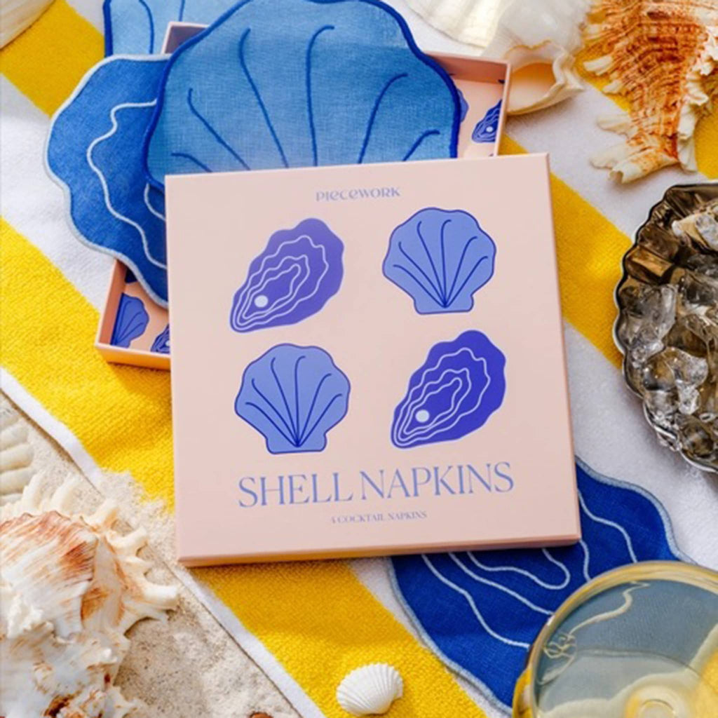 Piecework Linen Shell-Shaped Cocktail Napkins with pink box packaging on beach towel background with napkins and shells.