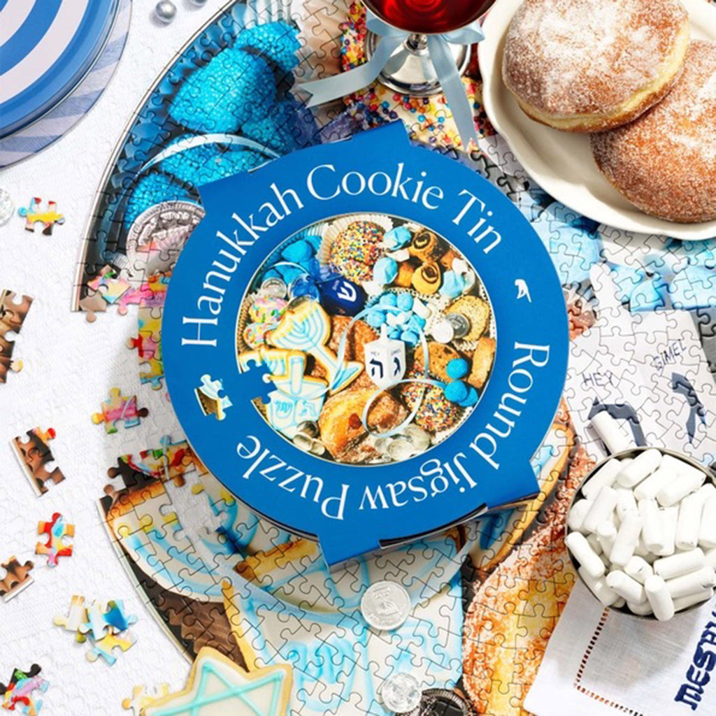 Piecework Puzzles 750 Piece Hanukkah Cookie Tin Round Jigsaw Puzzle in tin packaging with blue sleeve that has the puzzle image on it. Shown on top of the puzzle in progress with a plate of jelly dougnuts.