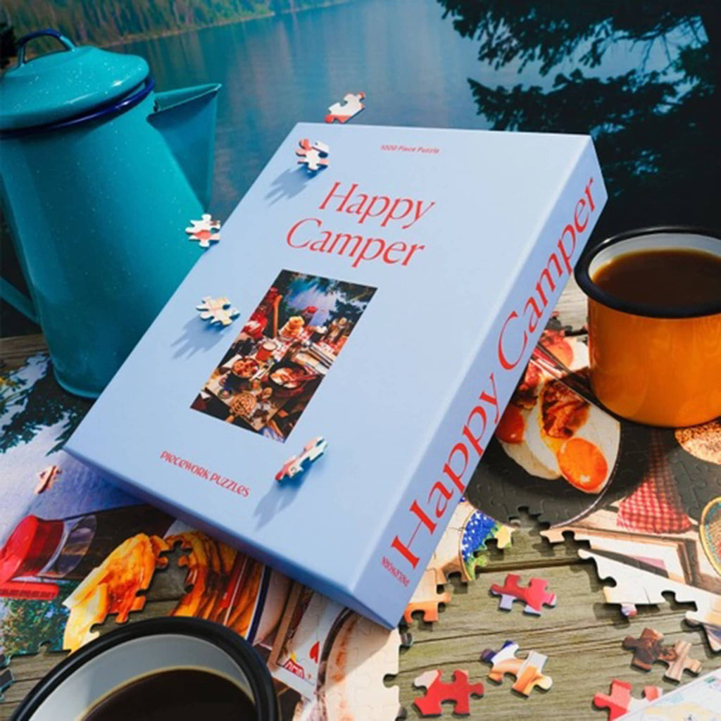 Puzzle box titled 'Happy Camper' with puzzle pieces and coffee cups on a table.