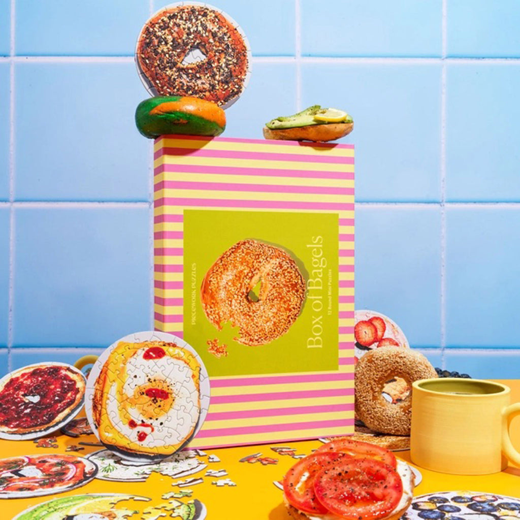 Piecework Puzzles Box of Bagels, set of 12 round mini puzzles in pink and lime green striped box packaging. Box is surrounded by mini puzzles in progress, real bagels with toppings, a yellow mug and yellow surface with a light blue tile wall behind.