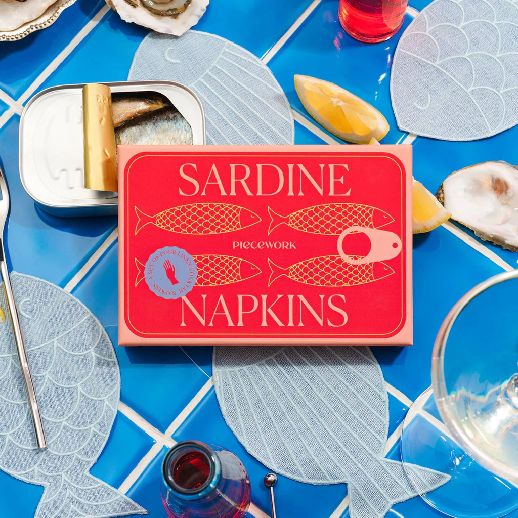 Piecework set of 4 sardine fish-shaped blue linen cocktail napkins in red box packaging, on blue tile background with sardines, oysters and lemons.