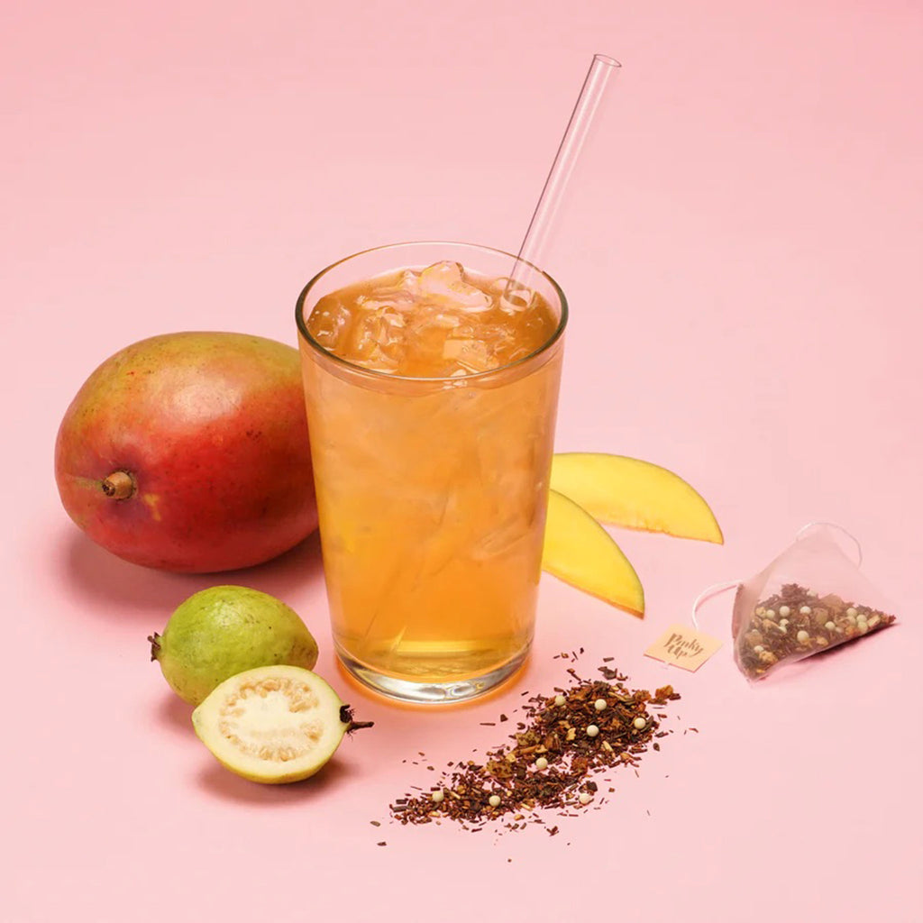 Pinky Up Mango Guava Boba Tea prepared in glass with straw with fruit and an open tea sachet beside it on a pink background.