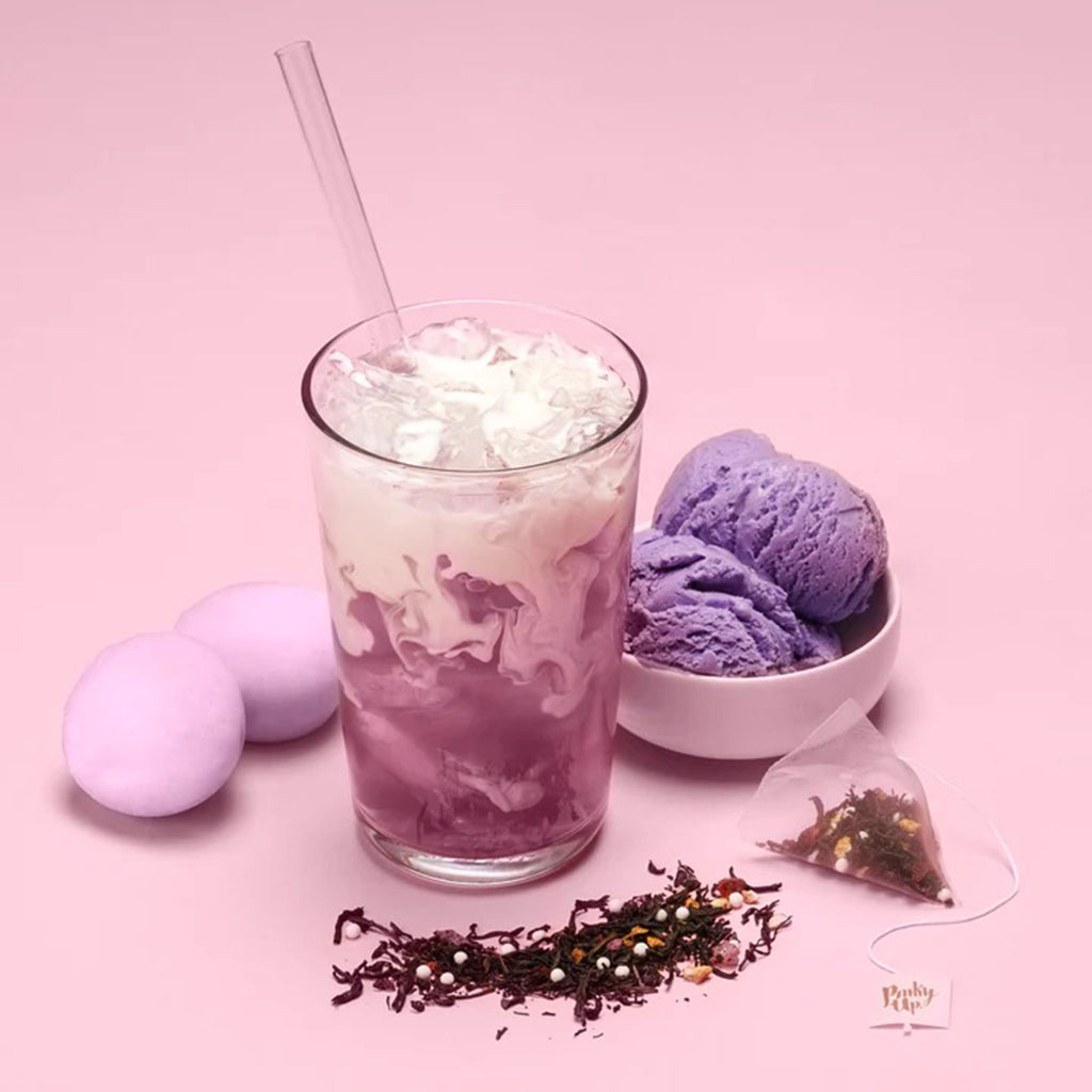 Pinky Up Mochi Ice Cream Boba Tea prepared with milk in a glass with straw with mochi, purple ice cream and an open tea sachet on a purple background.