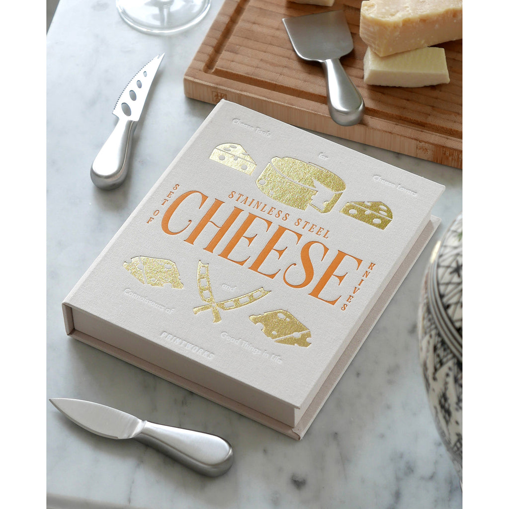 Printworks The Essentials Cheese Tools for Cheese Lovers, set of 3 stainless steel cheese knives with beige book box packaging, a serving board and cheese on a marble surface.
