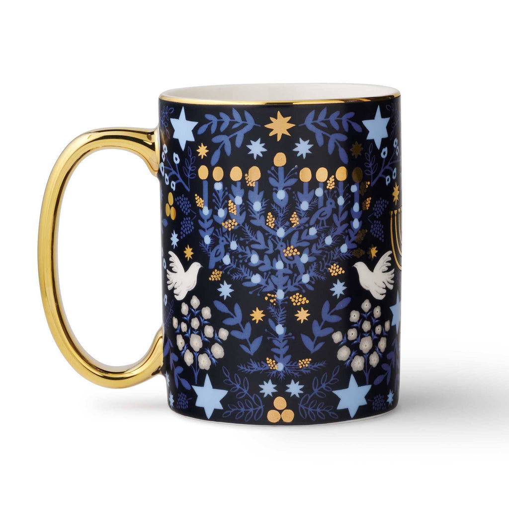 Mug with rifle paper laurel menorah pattern featuring stars, laurel, and birds on a navy background with gold rim and handle on the left, on a white background.