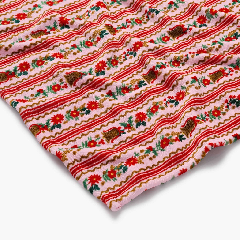 Pink fleece blanket with a festive pattern featuring red stripes, flowers, bells, and holly, corner detail view, on a white background.