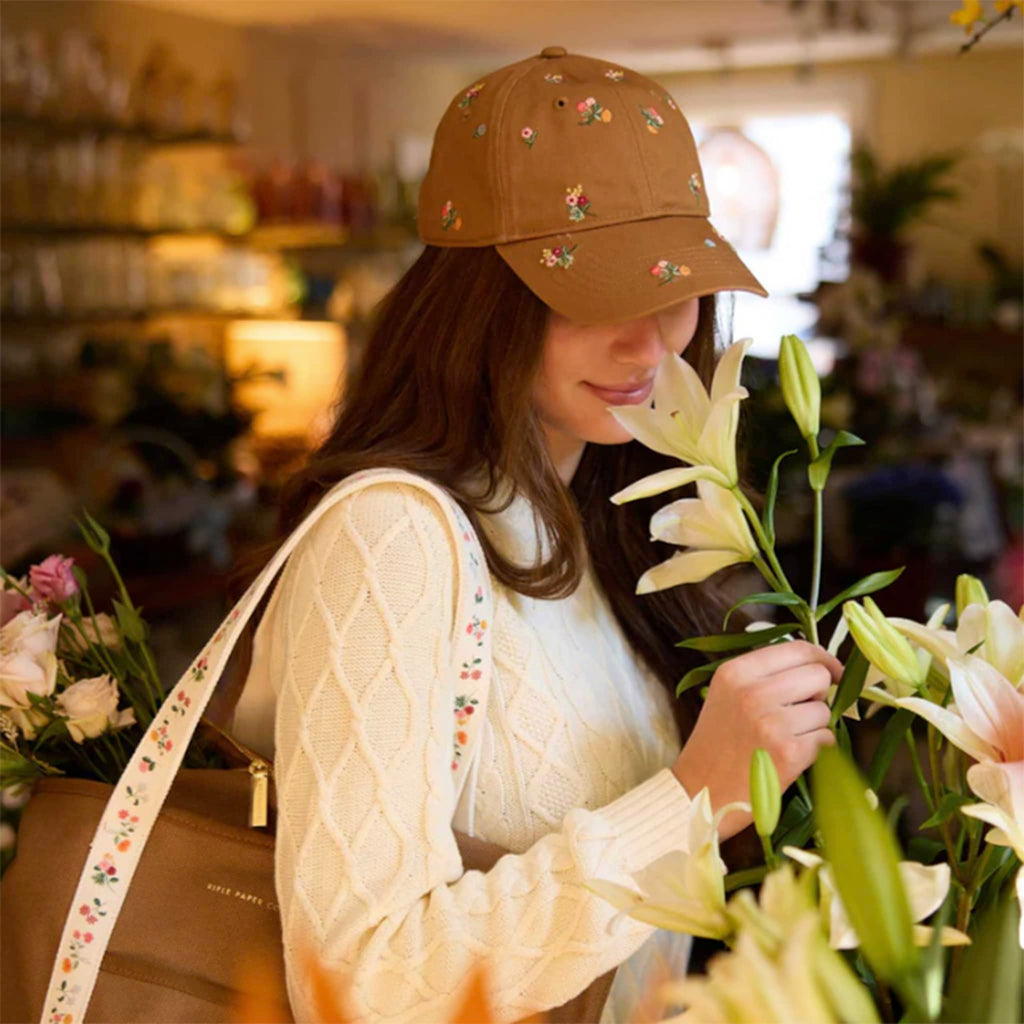 Woman wearing a brown floral baseball cap, holding flowers in a flower shop setting.
