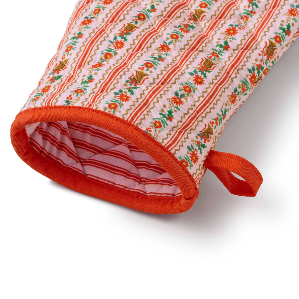 Decorative oven mitt with floral and bell pattern on the outside and pink and red striped print on the inside, detail of cuff, on a white background.