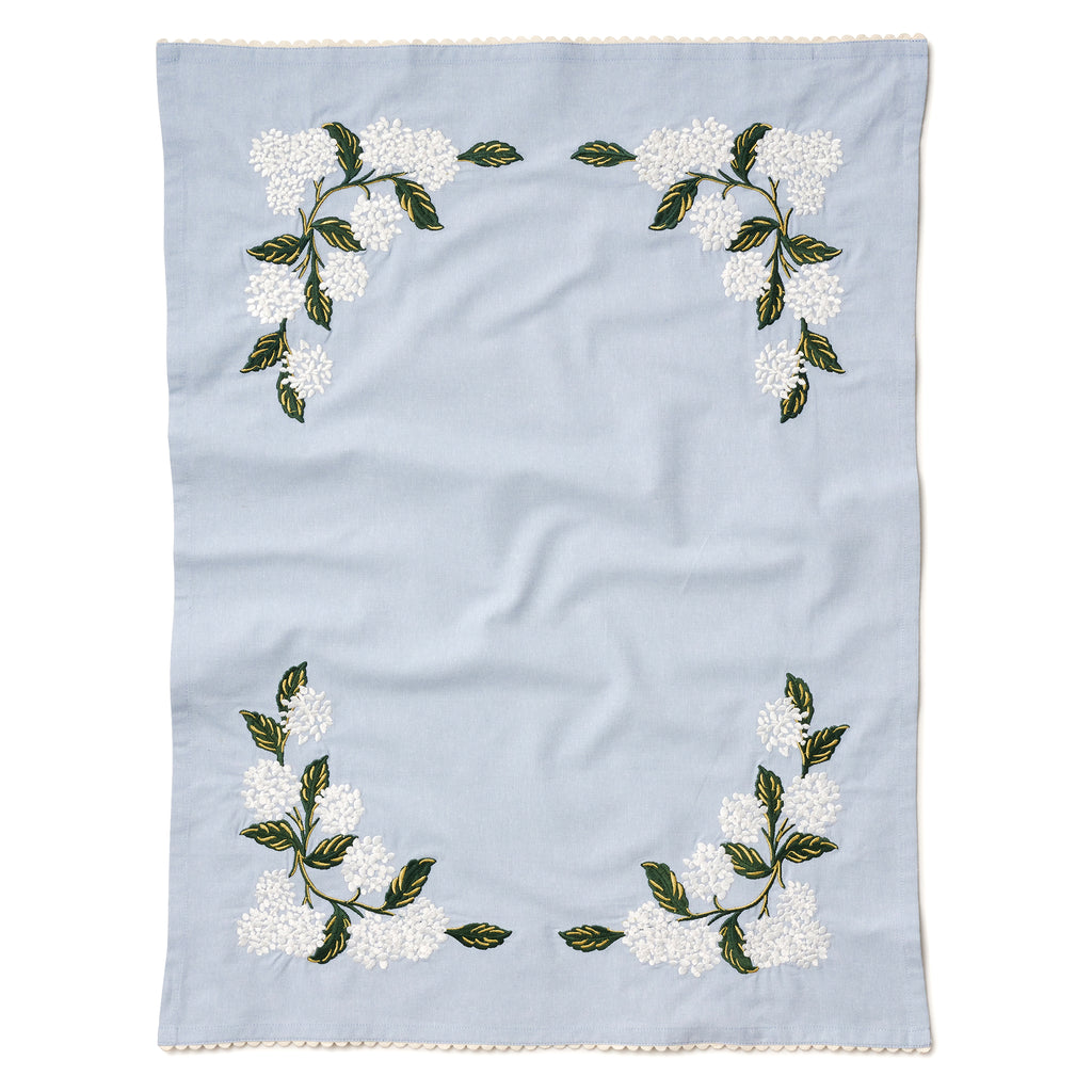 Rifle Paper Co. light blue tea towel with white hydrangea print embroidery  and white ric-rac edging, lying flat.