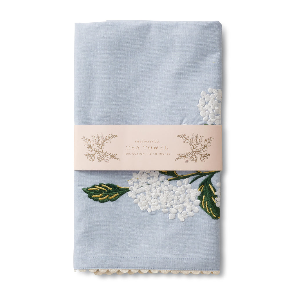 Rifle Paper Co. light blue tea towel with white hydrangea print embroidery  and white ric-rac edging, folded in belly band packaging.