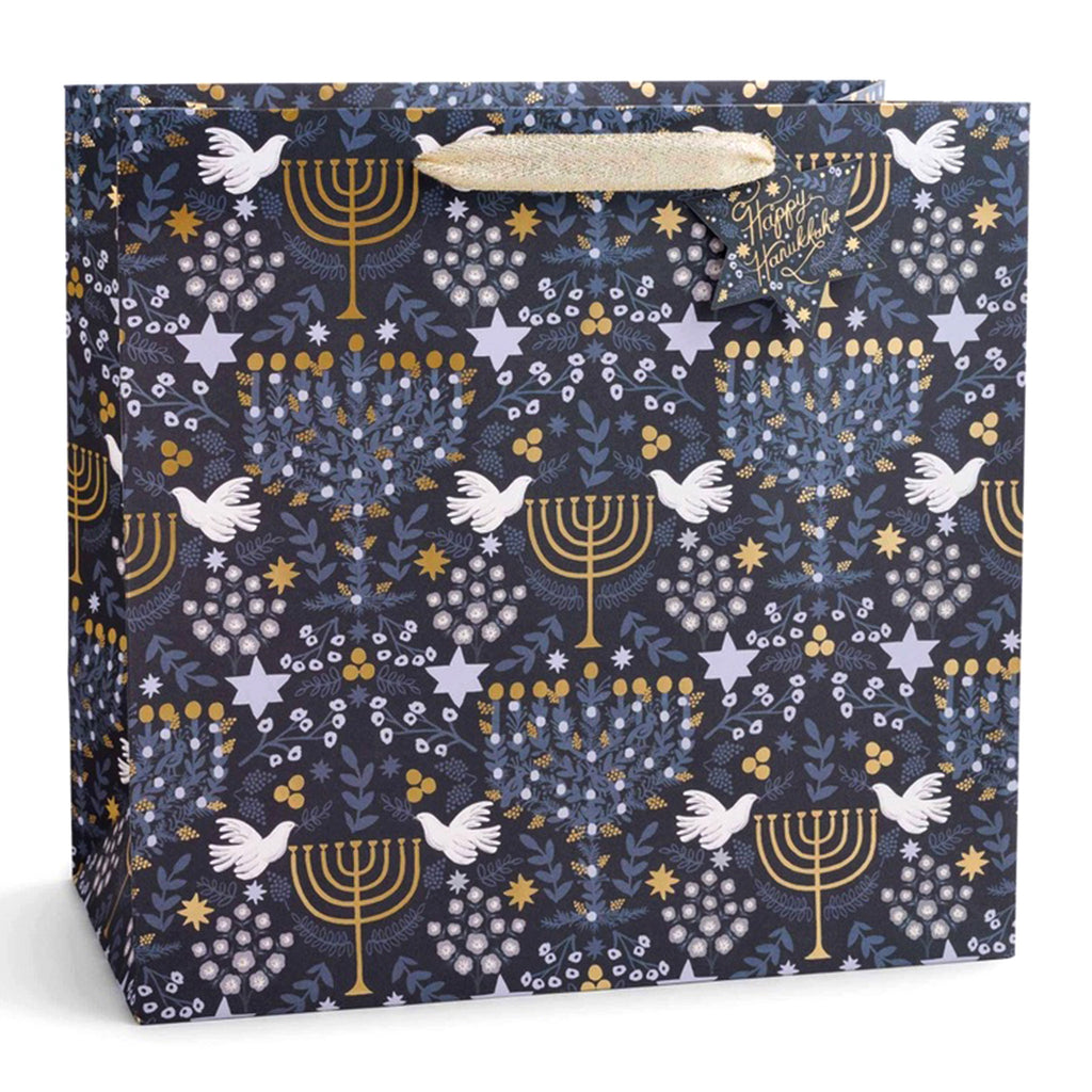 Rifle Paper Co. Laurel Menorah Hanukkah Large Gift Bag, front view. Navy blue bag with gold menorahs, white doves, leaves and Star of Davids in shades of blue with ivory cotton ribbon handles and a Star of David "to" and  "from" gift tag that says “Happy Hanukkah” in gold script lettering.