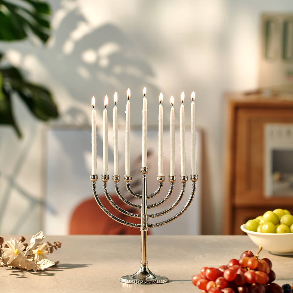 Silver Menorah with white lit candles on a table with fruits and flowers.
