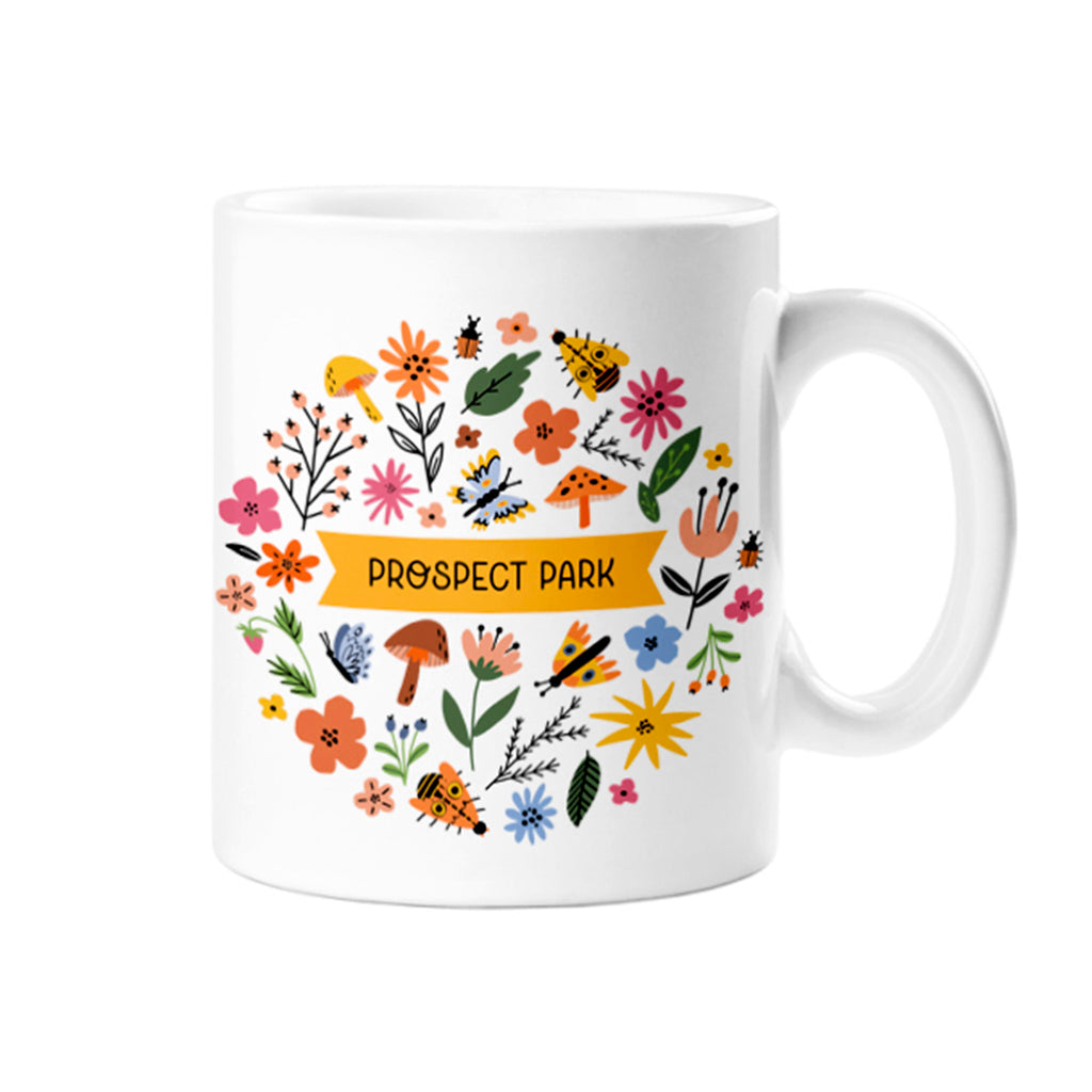 Rock Scissor Paper white ceramic mug with "Prospect Park" on a golden ribbon surrounded by flowers, bugs, leaves and mushrooms.