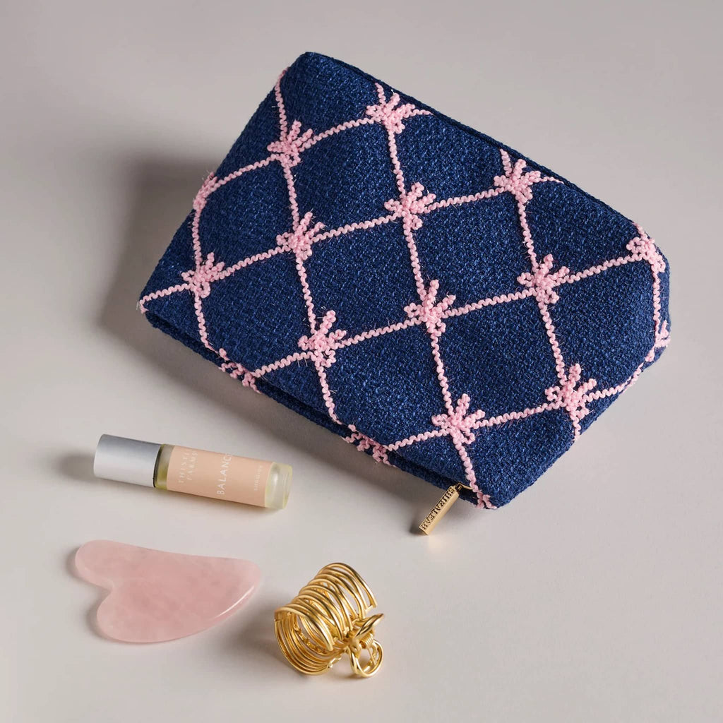 Navy clutch with pink bow pattern with a perfume bottle, lip balm, and gold hair clip on a light gray background.