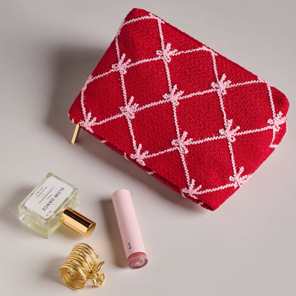 Red clutch with pink bow pattern with a perfume bottle, lip balm, and gold hair clip on a light gray background.