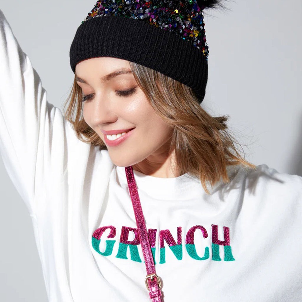 Shiraleah white sweatshirt with classic round neck and banded cuffs and hem and "GRINCH" in bright pink sequin and green sherpa bold lettering, shown on model with black sequin knit hat on.