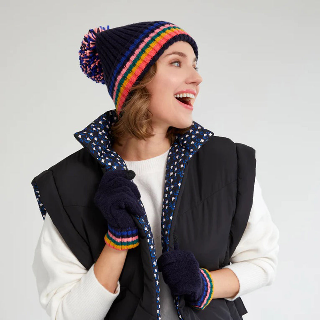 Shiraleah Ronen Knit Winter Hat in navy with colorful stripes and a big navy, blue and pink pom pom on top. Shown on model with matching Ronen gloves, a white sweater and a black puffy vest.