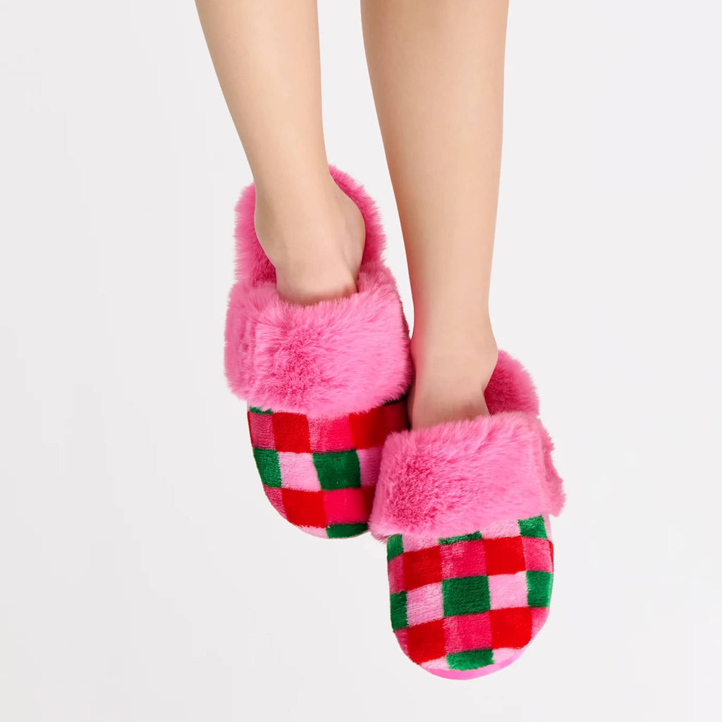 Pair of pink fuzzy slippers with pink, red and green checkered front on a person's feet, on a white background.