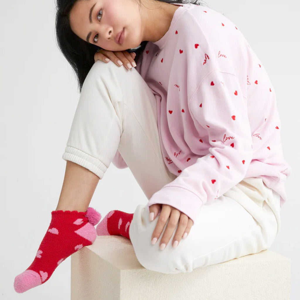 Shiraleah Hearts Home Socks, red plush socks with pink toe, heel and pompom at the ankle with pink heart pattern and rubber grip dots on the bottom. A model is wearing the socks along with white sweatpants and a pink sweatshirt, sitting on a white cube.