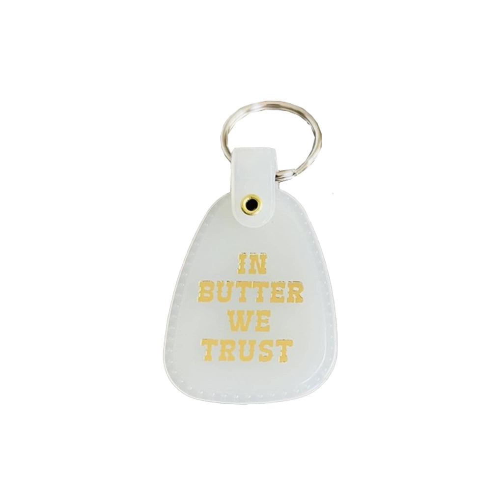 White saddle keychain with 'IN BUTTER WE TRUST' in gold text on a white background.