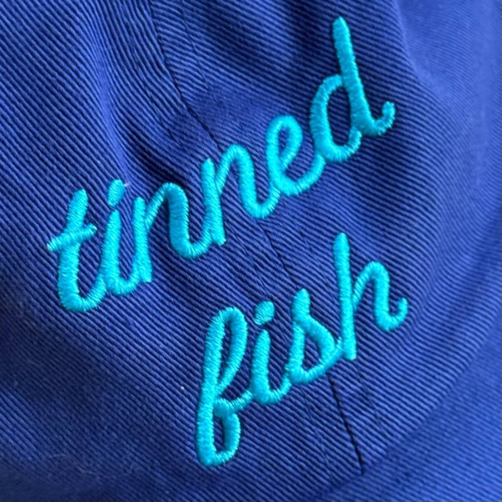 Bright blue cap with 'tinned fish' embroidered in turquoise, detail.