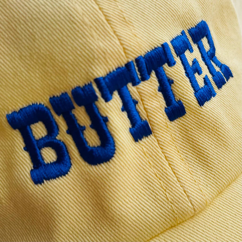 Silver Spider soft yellow unisex baseball cap with "butter" in blue embroidered letters, detail of embroidery.