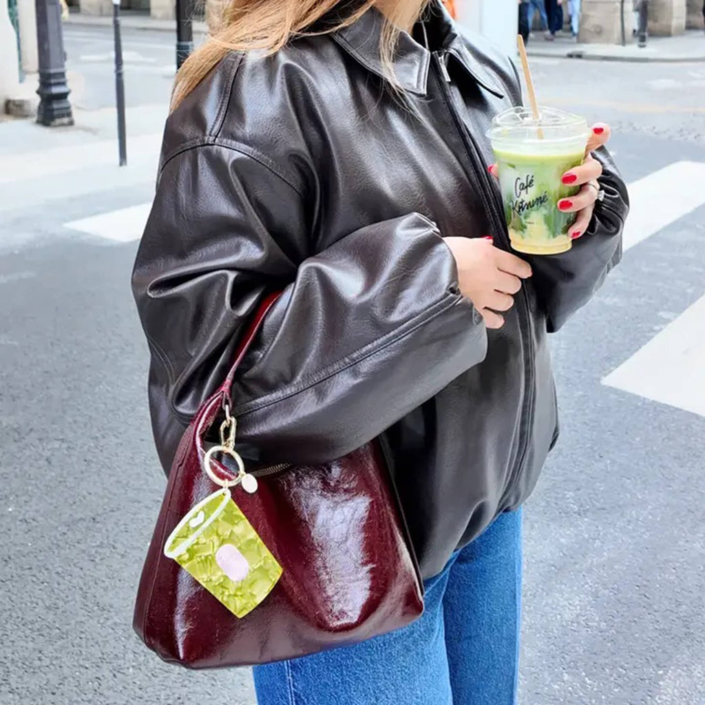 Person holding a cup of matcha with a burgundy bag and a gold tone keychain with green cup-shaped charm, on a city street.