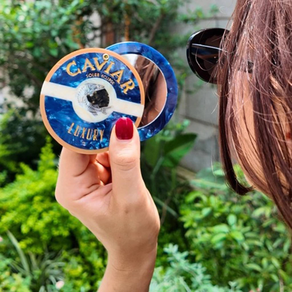 Person holding the Solar Eclipse compact pocket mirror with caviar tin design, with greenery in the background.