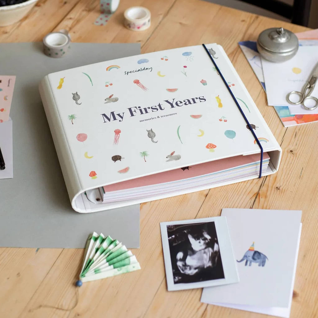 Memory book titled 'My First Years' on a wooden table with photos and stationery items.