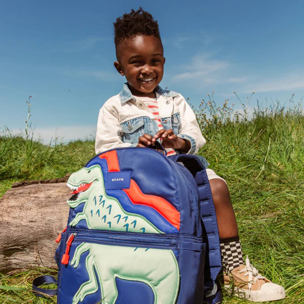 State Bags Kane Kids Travel Backpack in Dino, child holding it for scale in outdoor setting.