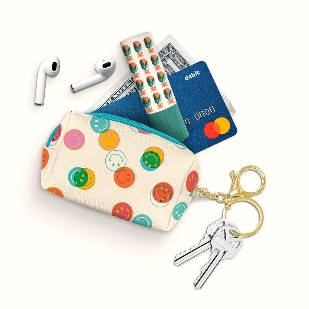 Studio Oh! Smiley Trails Keychain Zipper Pouch with colorful smiley faces on a cream color coated canvas pouch with turquoise zipper and gold tone key ring. Overhead view with airpods, lipbalm, money and a debit card spilling out of the pouch.