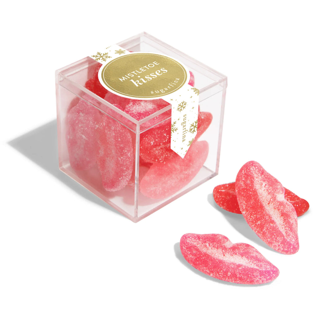 Sugarfina Mistletoe Kisses Sugar Lips red and pink lip shaped holiday gummy candy in small clear acrylic candy cube with a gold circular label on the top and a gold snowflake pattern on a white ribbon sticker. 3 pieces of lip shaped gummy candy are in front of the cube.