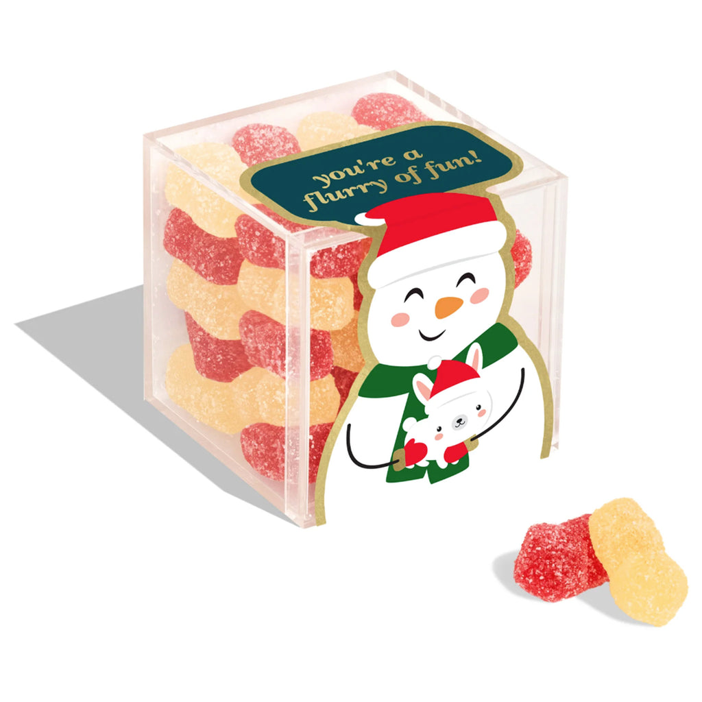 Sugarfina Snow Buddies snowmen shaped holiday gummy candy in small clear candy cube with a snowman holding a white dog on the front and "you're a flurry of fun" on the top in gold lettering on a green background. A red and a pale yellow snowman gummy are sitting in front of the cube.