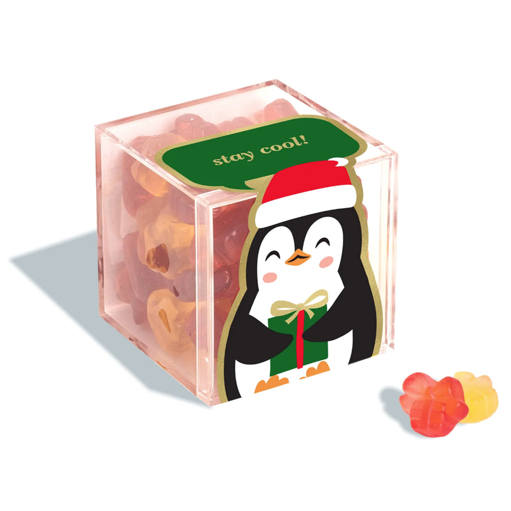 Sugarfina Penguin Presents holiday gummy candy in small clear acrylic candy cube with a penguin holding a gift on the front and "stay cool" in gold lettering on a green background on the top. A red and a yellow present shaped gummy are in front of the cube.