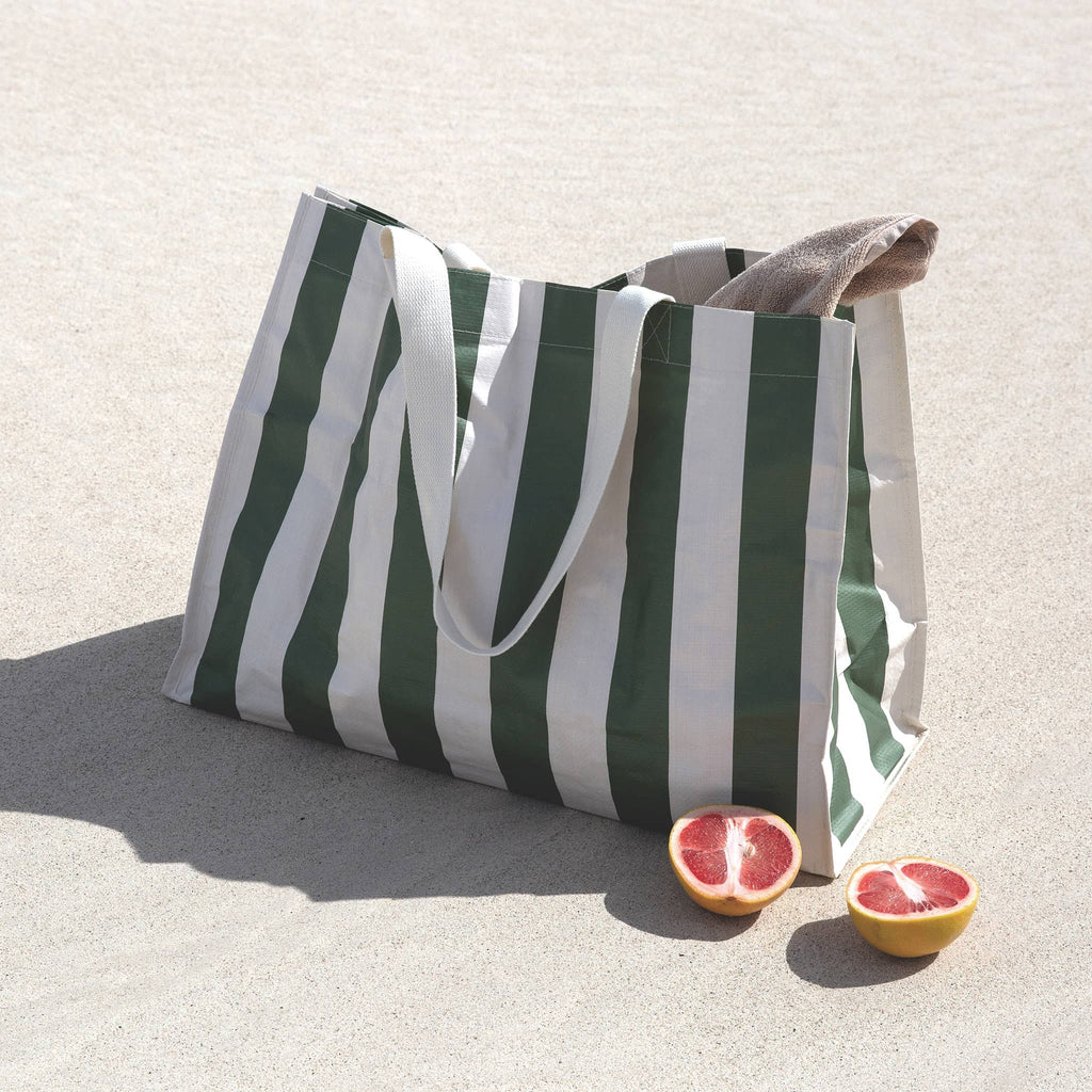 Sunnylife Carryall Beach Bag in The Vacay Olive Stripe, front angle view on a beach with towel inside and a blood orange sliced in half.