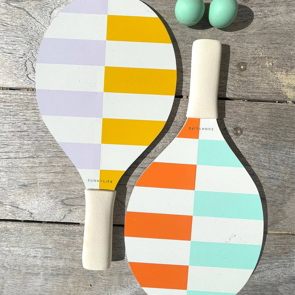 Sunnylife Rio Multi Beach Bats with colorful stripes, white soft grips and aqua balls, on wood surface.