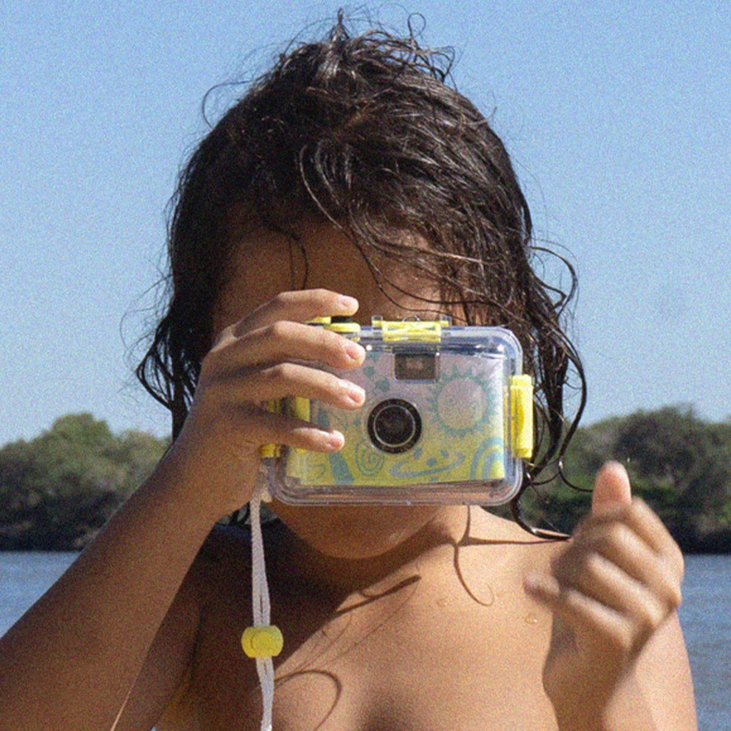 Sunnylife Sea Kids 35mm waterproof underwater camera with green illustrations on an ombre yellow to white background, with child taking a picture with it.