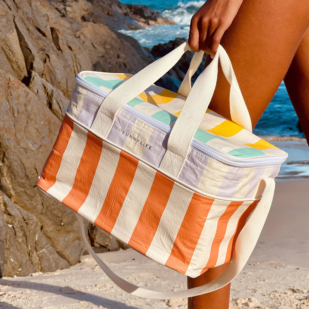 Sunnylife Cooler Bag in Rio Sun with blue, yellow, lilac, orange and cream maxi stripes, being carried on the beach.